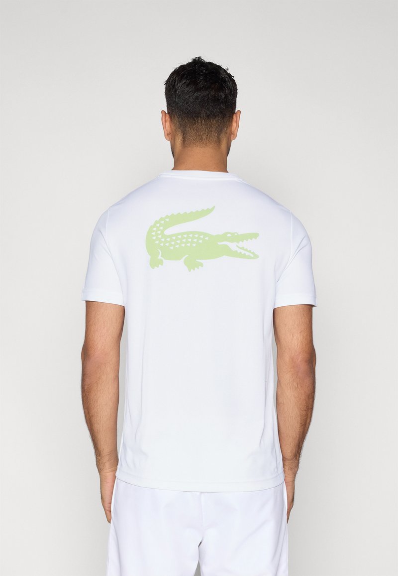 Biała koszulka z krótkim rękawem wykonana z gładkiej tkaniny, z jasnym zielonym nadrukiem krokodyla na górnej części pleców. Klasyczny, okrągły dekolt.