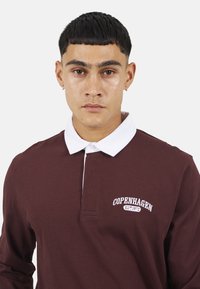 Polo a maniche lunghe bordeaux con colletto bianco e logo ricamato "COPENHAGEN EST. 1972" sul lato sinistro. Materiale in cotone.