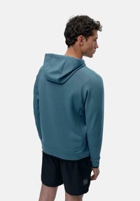 Kapucinska majica v teal tkanini, ki ima prilagojen kroj in kenguru žep, v kombinaciji z črnimi kratkimi hlačami, ki so vidne na dnu.