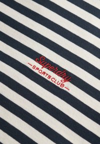 Granatowo-białe paski na tkaninie o gładkiej fakturze. Na środku znajduje się czerwony haftowany napis "Superdry" oraz "SPORTS CLUB".