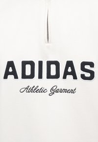 Indumento sportivo bianco con colletto a metà zip. Logo "ADIDAS" ricamato in nero con "Athletic Garment" in corsivo sottostante. Texture liscia.