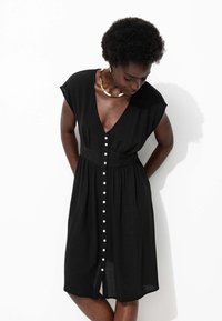 Robe noire jusqu'aux genoux avec un décolleté en V profond, des manches courtes et des détails de boutons blancs sur le devant. Fabriquée en tissu léger et texturé.