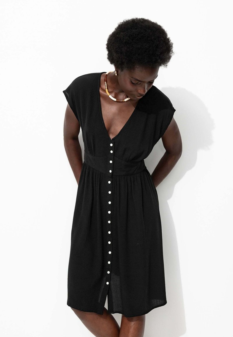 Robe noire jusqu'aux genoux avec un décolleté en V profond, des manches courtes et des détails de boutons blancs sur le devant. Fabriquée en tissu léger et texturé.