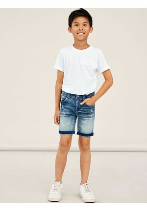 T-shirt blanc à manches courtes avec poche, associé à un short en denim déchiré, présentant un ourlet retroussé et un dégradé de bleu clair à foncé. Baskets blanches.