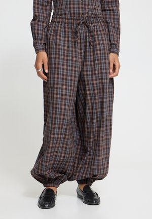 Personne portant un pantalon ample à carreaux marron et bleu avec des poignets élastiques et des chaussures noires à enfiler, debout devant un fond blanc.