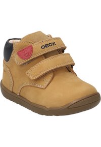 Zapato de ante para niño pequeño en color marrón claro con dos correas de velcro, suela de goma marrón y acento negro en el talón. Presenta un parche de logo rojo en el lado exterior.