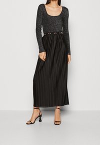Černá plisovaná maxi sukně v kombinaci s třpytivým černým dlouhým rukávem. Oblečení má pás s logem značky a sandály na podpatku s otevřenou špičkou.