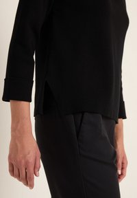 Svart stickad långärmad topp med sidofickor, bärs över mörka smala byxor. Ärmsluten är uppvikta, vilket visar en slät textur och en strukturerad design.