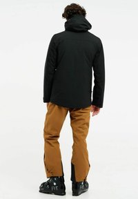 Schwarze wasserdichte Jacke mit Kapuze und braune Hose. Die Hose hat schwarze Akzente und Reißverschlüsse am Knöchel. Schwarze Skischuhe mit blauen Details.