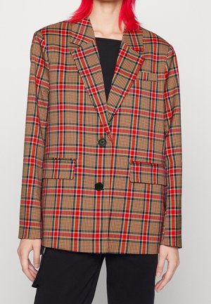 Persoon met rood haar die een rood, zwart en beige geruit colbert draagt over een zwart shirt en zwarte broek tegen een neutrale achtergrond.