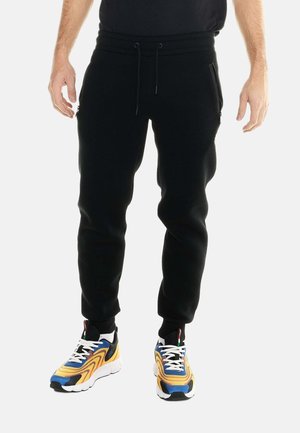 Uomo che indossa pantaloni da jogging neri con tasche con zip e sneaker multicolore gialle, blu e bianche, che sta in piedi su uno sfondo bianco.