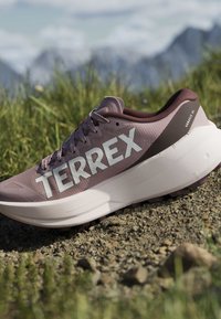 Ανοιχτό μωβ παπούτσι τρεξίματος Adidas Terrex με παχιά λευκή σόλα σε χαλίκι με χόρτο και βουνά στο φόντο.