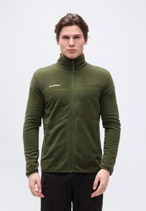 Młody mężczyzna w zielonej polarowej kurtce Mammut z zamkiem i czarnych spodniach, stojący na tle jednolitego jasnego tła.