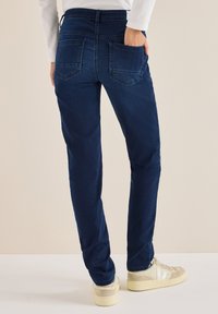 Vaqueros azul marino de corte slim, con una cintura alta, dos bolsillos traseros y una textura sutil. Combinados con zapatillas de color claro.