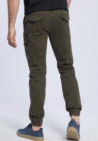 Oliwkowe spodnie cargo z elastycznymi ściągaczami, wieloma bocznymi kieszeniami oraz miękkim, fakturowanym materiałem, zestawione z niebieskimi trampkami z nakrapianą podeszwą.