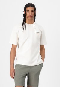 Cremefarvet bomulds t-shirt med korte ærmer, prydet med et McLaren-logo i sort og orange. Sammen med teksturerede olivengrønne shorts.