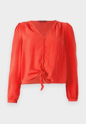 Sarkana garā piedurkņu blūze ar V-veida kakla izgriezumu un frontālu pogu aizdari. Iekļauj jostu viduklī un ir izgatavota no mīksta, teksturēta auduma.