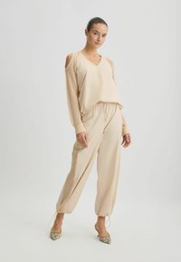 Beige loungewear set met een V-hals top met mesh mouwen en broeken met een trekkoord en cuffed enkels, aangevuld met patroon hoge hakken.