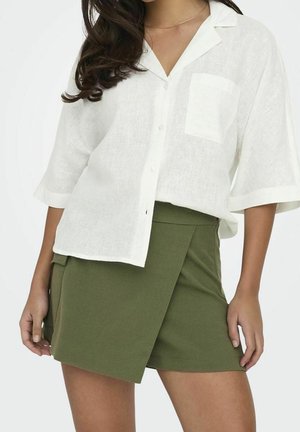 Femme portant une chemise blanche boutonnée avec une poche et une jupe portefeuille vert olive, debout devant un fond uni.