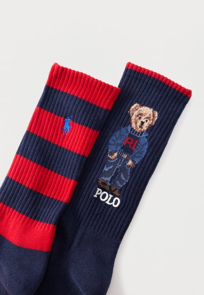 Κάλτσες με ρίγες σε ναυτικό και κόκκινο χρώμα, η μία με μπλε λογότυπο παίκτη πόλο και η άλλη με αρκούδα που φοράει μπλε πουλόβερ και τζιν, με το κείμενο "POLO".