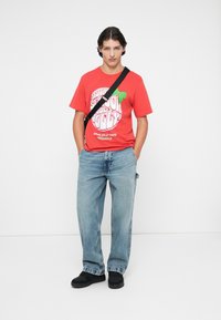 Crvena pamučna majica s grafičkim natpisom i zelenim naglaskom, uparena s laganim plavim trapericama i crnim natikačama. Torba s podesivim remenom.