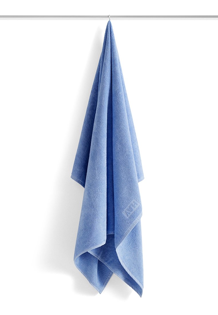 Blauwe handdoek hangend aan een zilveren stang tegen een witte achtergrond, met een klein, textuurachtig merklabel nabij de onderrand.