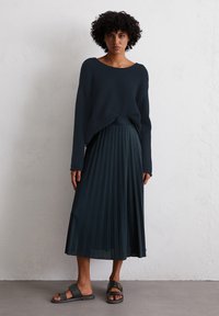 La modella è in piedi contro un muro testurizzato, indossando un maglione blu navy dal taglio morbido, una gonna plissettata blu navy e sandali neri. L'espressione è neutra.