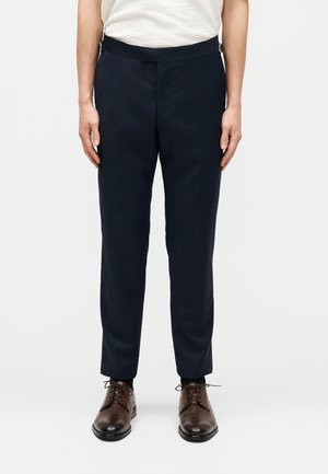 Mann trägt schwarze maßgeschneiderte Hose und dunkelbraune Lederschuhe, steht vor schlichtem weißem Hintergrund.