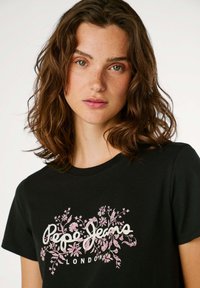 Melna kokvilnas T-kreklā ar rozā ziedu rakstiem un balto uzrakstu "Pepe Jeans London", ar apaļu kaklu un īsām piedurknēm.
