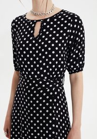 Robe noire à pois comportant un décolleté en trou de serrure, des manches courtes bouffantes et un lien à la taille. Fabriquée dans un tissu léger avec une texture lisse.