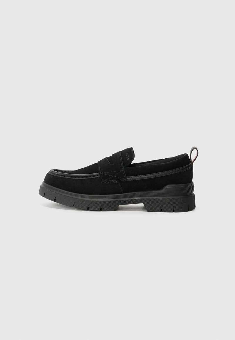 Sapatos slip-on pretos em camurça com uma sola de borracha preta espessa, apresentando uma correia superior e detalhes de costura contrastantes.