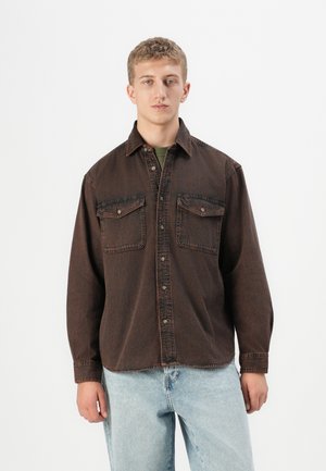 Young man with short hair wearing a dark brown buttoned denim shirt and light blue jeans, standing against a plain white background.