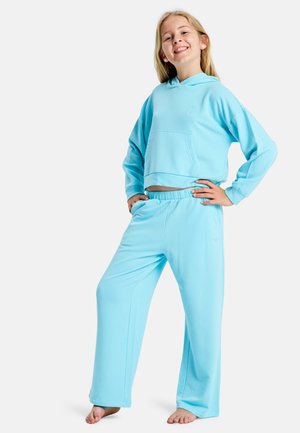 Lächelndes Mädchen, barfuß stehend, in hellblauem Hoodie und passenden Jogginghosen mit Händen in der Hüfte vor einem einfarbigen weißen Hintergrund.