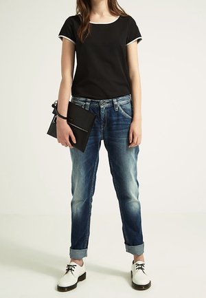 Top negro de manga corta con ribete blanco, jeans azules enrollados, zapatos blancos con cordones negros, sosteniendo un bolso de mano negro.