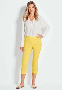 Gele cropped pants met een gladde textuur, gecombineerd met een witte blouse met lange mouwen. Model staat tegen een effen achtergrond, handen op de heupen.