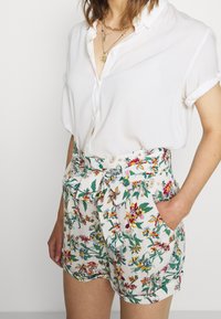 Shorts fleuris avec une base blanche, ornés de motifs floraux colorés en rouge, jaune et bleu. Les shorts sont dotés d'une ceinture à nouer et de poches latérales.
