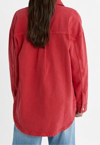 Camicia in denim rosso con vestibilità rilassata, maniche lunghe e polsini con bottoni. Presenta un orlo curvo e dettagli di cuciture a vista. Design casual.