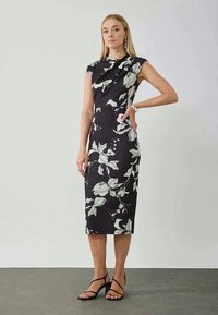 Zwarte aansluitende jurk met wit bloemenpatroon, met korte mouwen en een hoge hals, tot halverwege de kuit, gecombineerd met zwarte sandalen met bandjes.