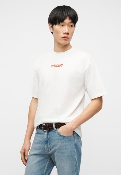 Jeune homme aux cheveux noirs portant un T-shirt blanc avec le texte « WRONG » et un jean bleu clair, une main dans la poche, regardant vers la droite.
