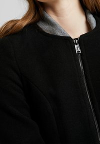 Jongeman in een zwarte ritsjack over een grijze hoodie, waarop de rits- en kraagdetails zichtbaar zijn, met lange, bruine haren in het zicht.