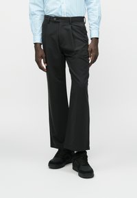 Pantaloni negri cu picioare largi, cu pliuri frontale, purtați cu o cămașă cu mânecă lungă în dungi albastre deschise și adidași negri. Textura materialului este netedă.