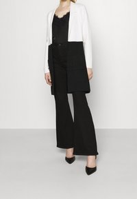 Black and white long cardigan with pockets, paired with a black lace top and flared black pants. Black pointed-toe heels complete the look.