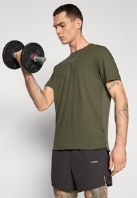 Oliven grønn treningstrøye med rund hals og logo, kombinert med svarte shorts. Holder en svart håndvekt i en treningsposisjon.