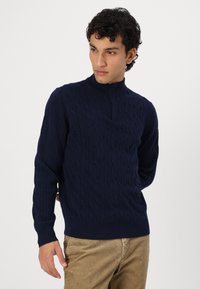 Granatowy sweter dzianinowy z wysokim kołnierzem, z teksturowanym wzorem w warkocze oraz ściągaczami przy mankietach i dole. Luźny krój, miękka tkanina.