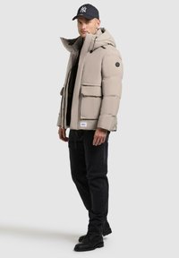 Beige puffer jacket with a hood, featuring two front pockets and a logo patch, paired with black jeans and black boots.