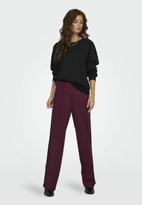 Zwarte oversized sweatshirt met lange mouwen, gecombineerd met bordeauxrode wijde broek met zijstrepen in het zwart. De outfit wordt compleet gemaakt met zwarte schoenen.