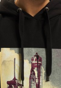 Fekete kapucnis pulóver texturált felülettel, elöl egy spray kanna és egy szerkezet grafikus nyomtatványával, megerősített zsinórvégekkel.