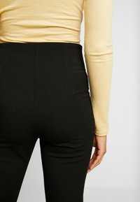 Svarta högmidjade byxor med en slät yta, tillsammans med en figurnära långärmad gul topp. Fokusera på midjan och bakifrån.