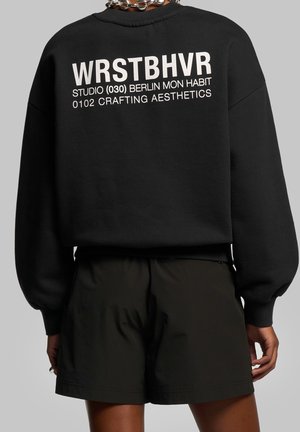 Sort sweatshirt med hvid tekst "WRSTBHVR STUDIO (030) BERLIN MON HABIT 0102 CRAFTING AESTHETICS" båret med sorte shorts og kædehalskæde.