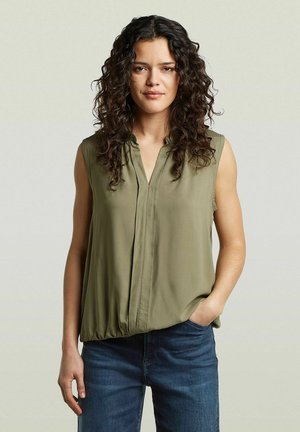 Femme aux longs cheveux bouclés foncés portant un chemisier sans manches vert olive et un jean bleu, debout avec une main dans sa poche.